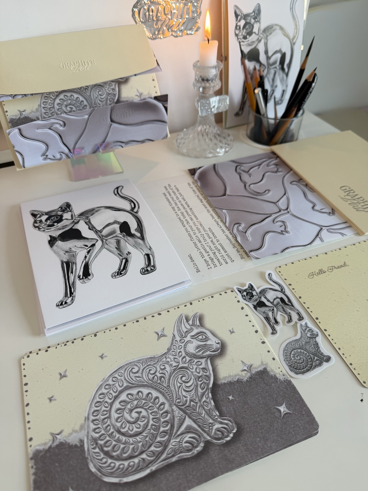 Graphite Post cat collection — full package flatlay on desk with print, postcard, stickers and letter beside a lit candle