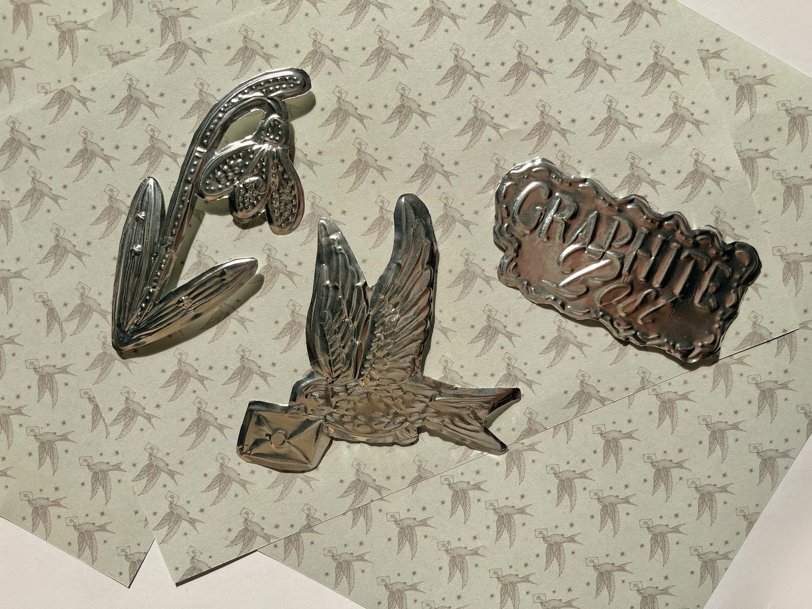 Premium metallic decorative objects from Graphite Post: swallow bird, snowdrop flower, and branded emblem on patterned paper
