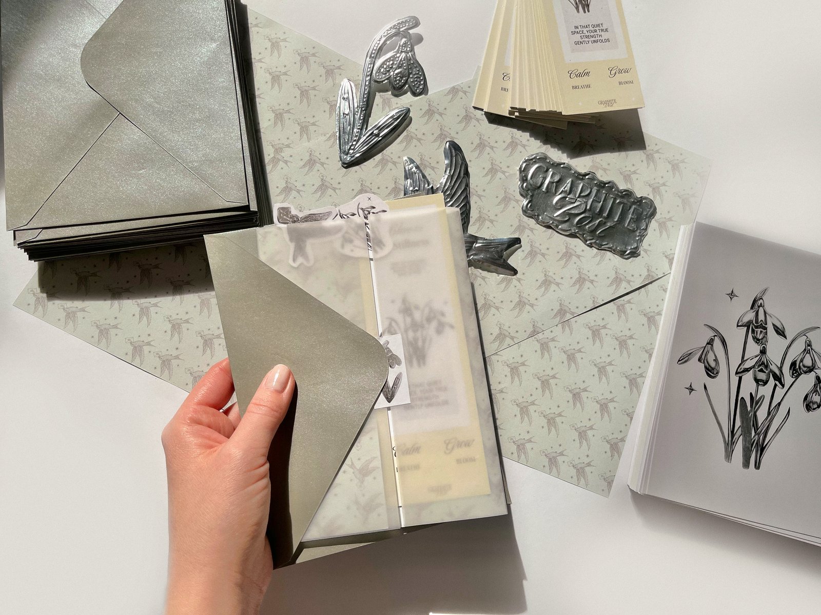Hand holding a silver envelope and a vellum-wrapped package above silver stickers and a snowdrop print on a white table