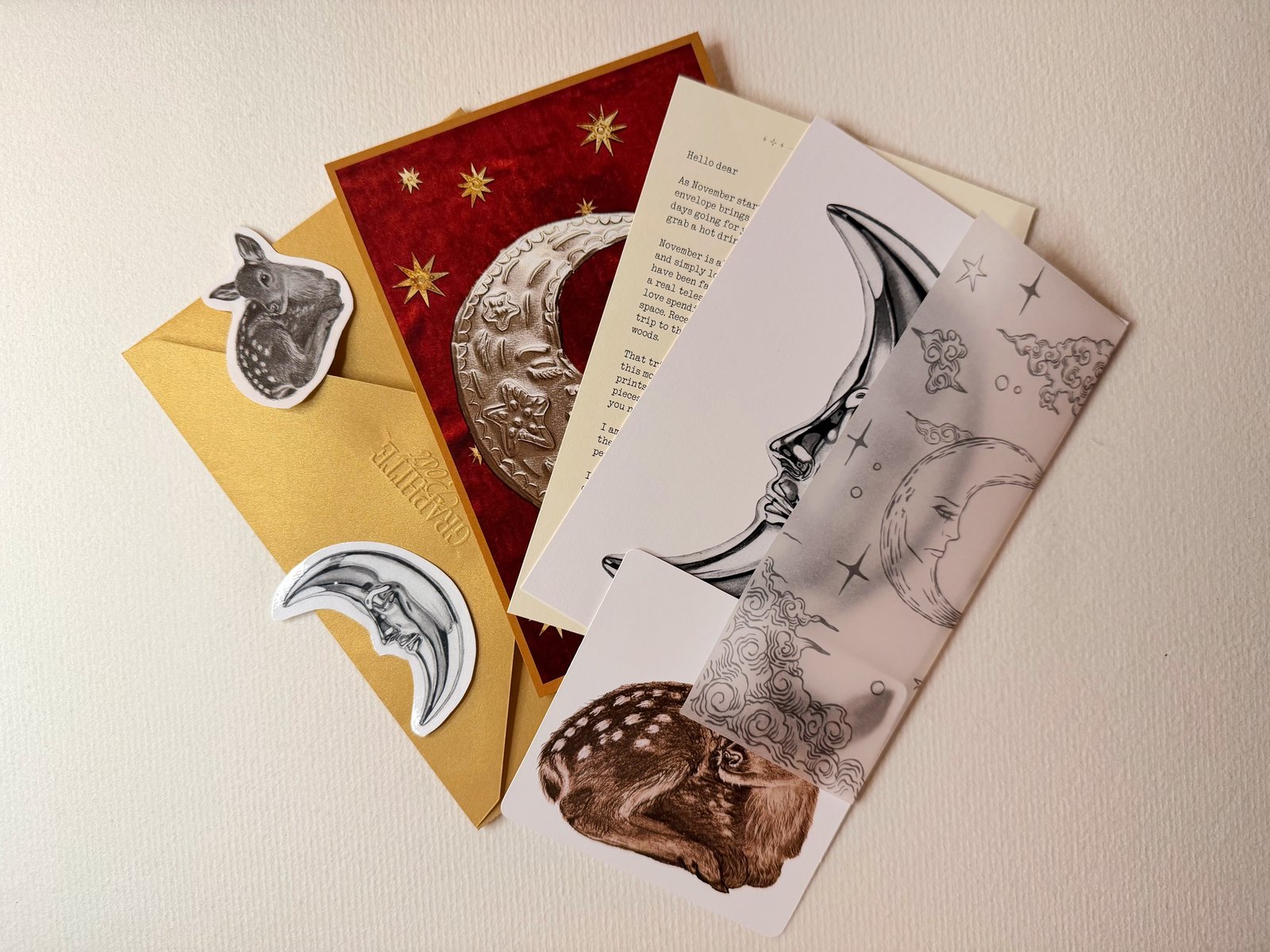 The contents of one envelope fanned out over the gold envelope on textured paper: a deep red collage print with an embossed silver moon, a typed letter, the moon print, vellum printed with moons and clouds and a small fawn print, with a fawn sticker and a moon sticker on the envelope