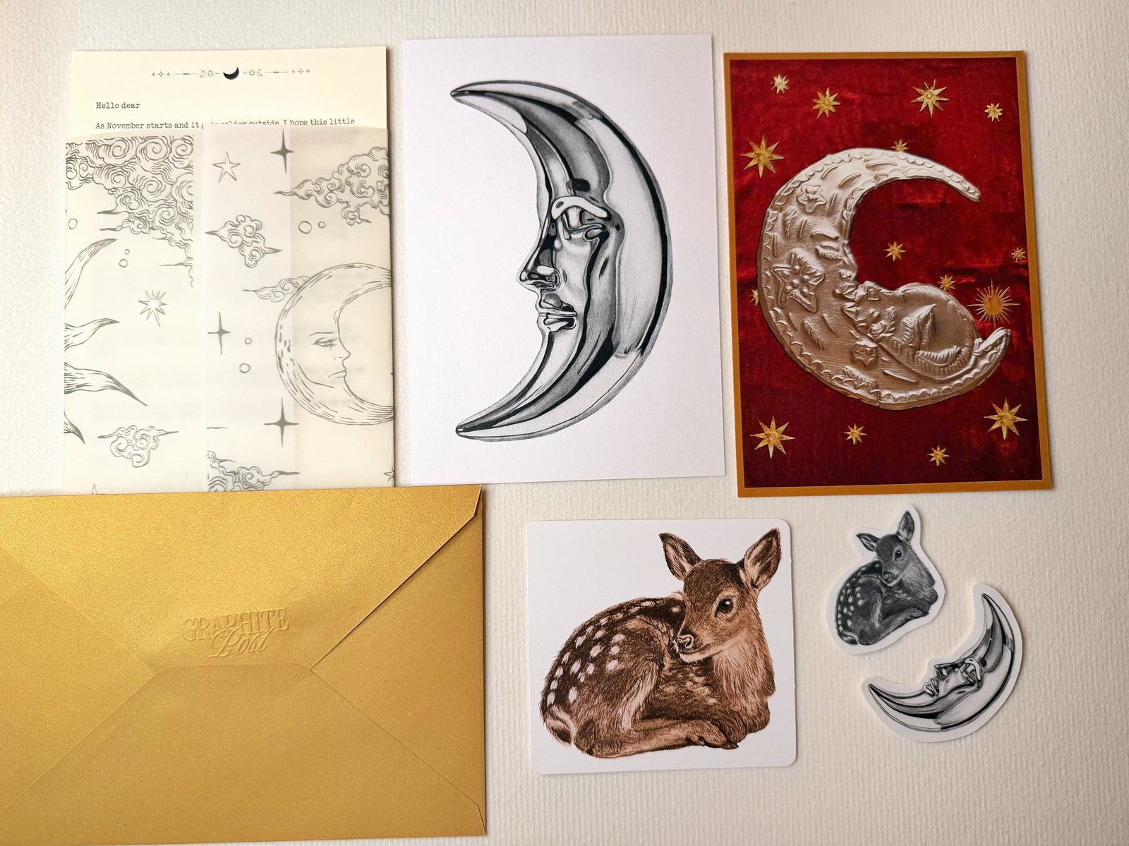 The contents of one envelope laid out on textured paper: a typed letter under vellum printed with moons and clouds, the moon print, a deep red collage print with an embossed silver moon and gold stars, a small square fawn print, a moon sticker and a fawn sticker, and the gold envelope