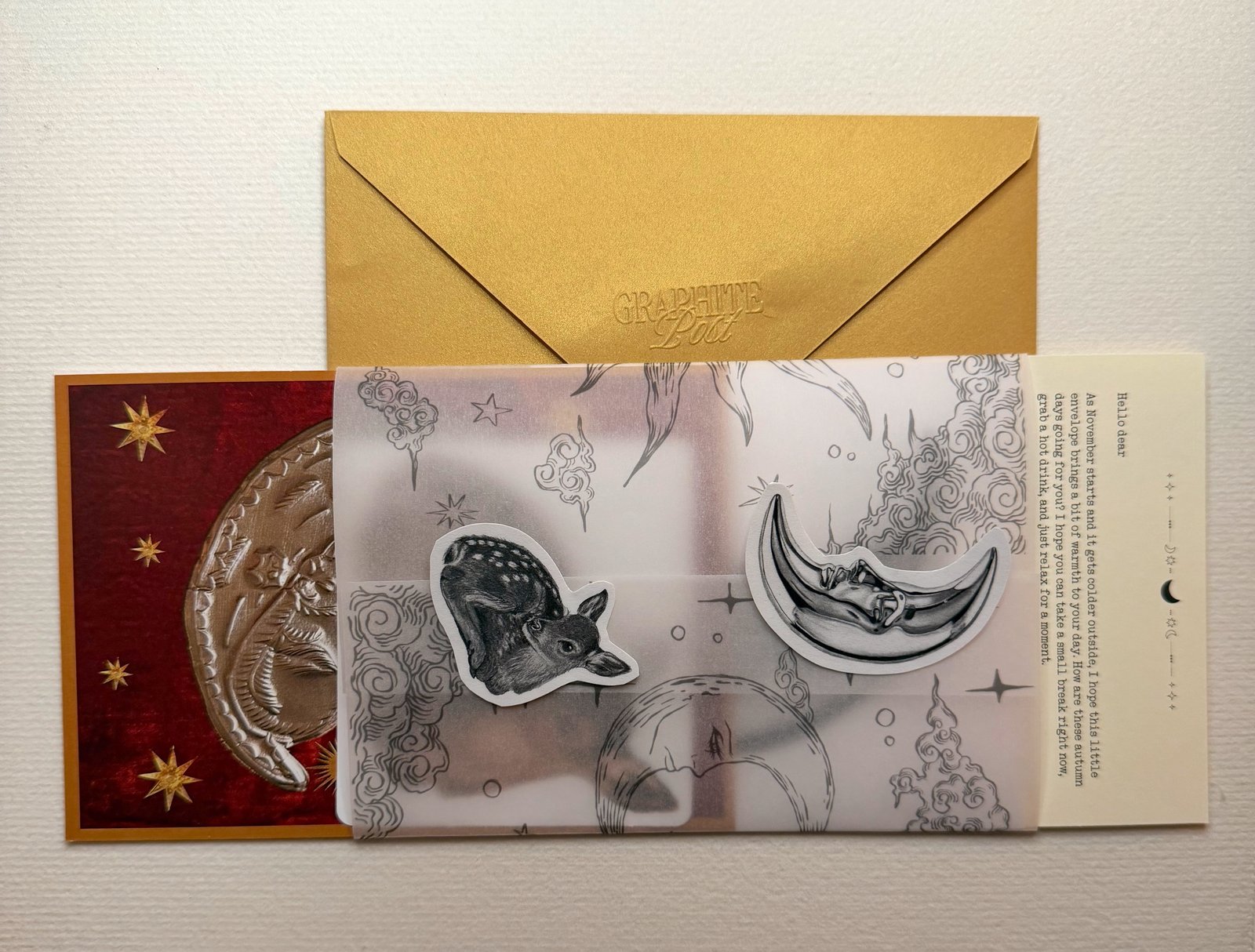 A closed gold envelope with the Graphite Post logo embossed on the flap, lying above vellum printed with moons and clouds that carries a fawn sticker and a moon sticker, with a deep red collage print at its left and a typed letter at its right, on textured paper