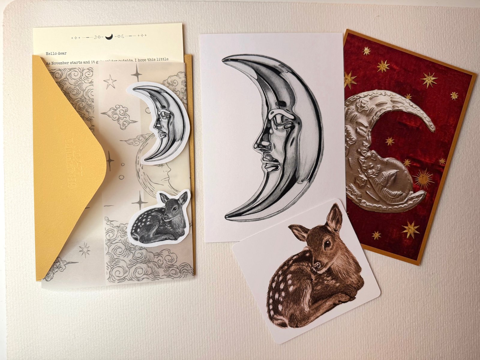 A gold envelope lying open on textured paper, with a typed letter and vellum printed with moons and clouds coming out of it, a moon sticker and a fawn sticker on the vellum, next to the moon print, a deep red collage print with an embossed silver crescent moon and gold stars, and a small square fawn print