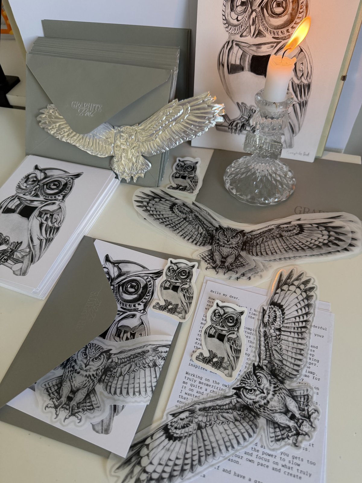 Owl prints, stacked grey envelopes and postcards, stickers, a silver owl ornament and a lit candle on a desk