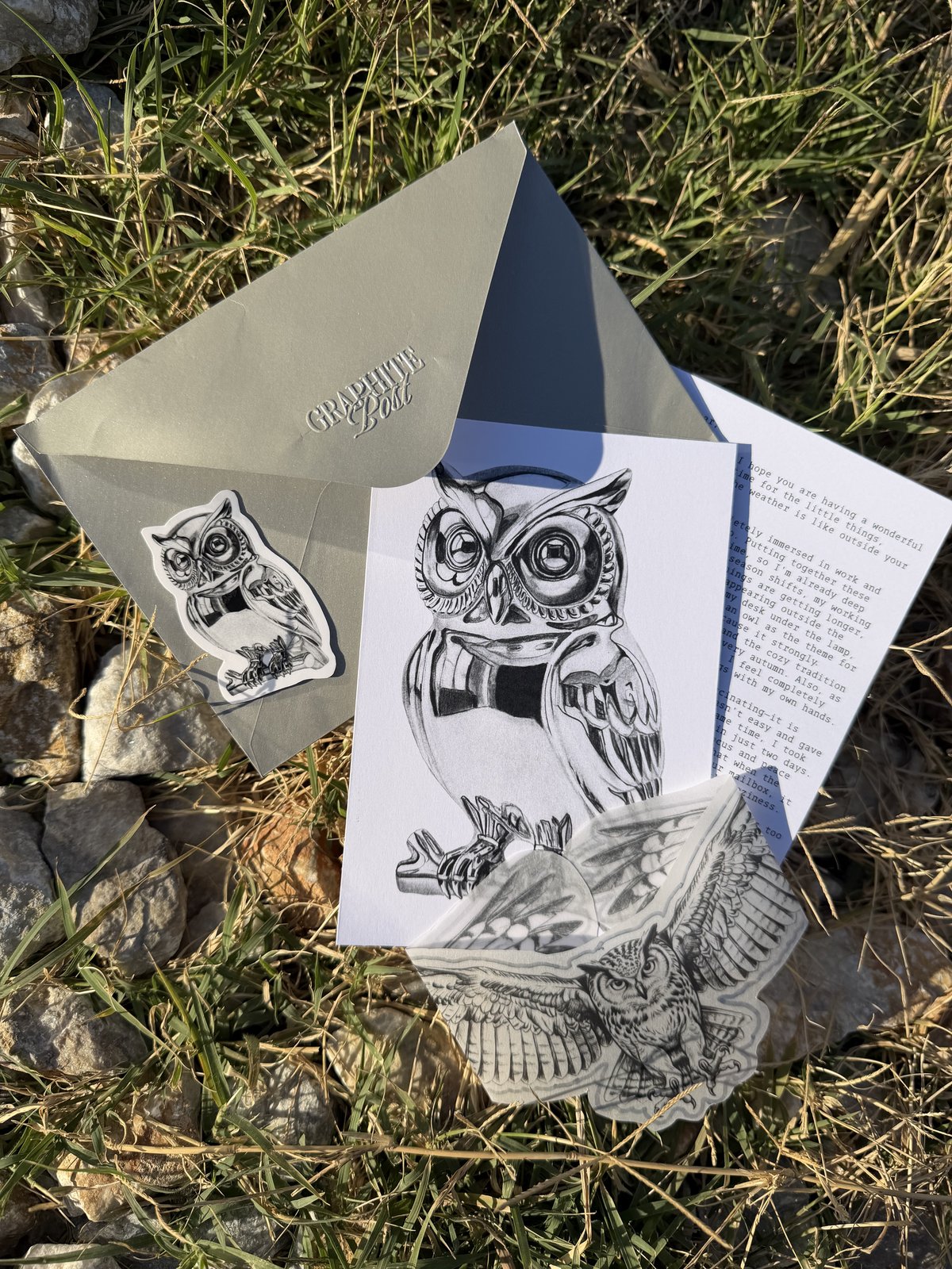 Owl collection, open gray envelope with the owl print, a typed letter and two owl stickers on grass and stones