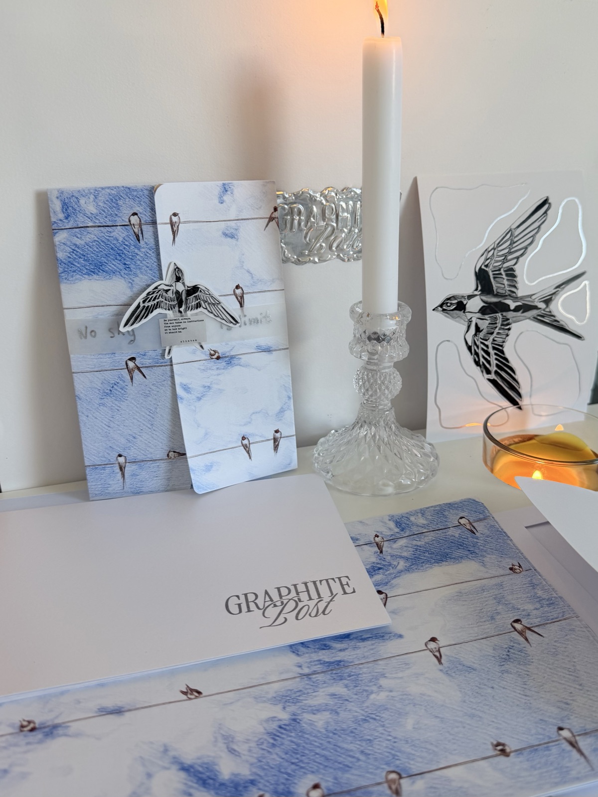 Graphite Post swallow collection — wire-and-birds cards and print beside a lit candle